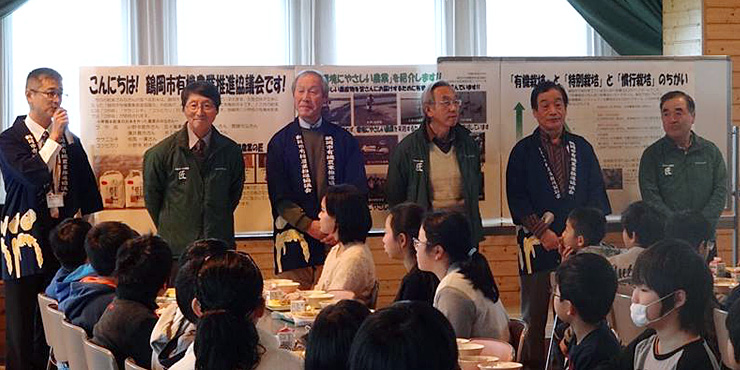 有機米給食を楽しむ小学生よ有機農業を説明する農家さんの画像